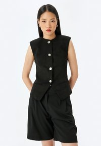 Zwarte mouwloze vest met een getailleerde snit, voorzien van zes zilveren knopen en twee voorzakken, gecombineerd met bijpassende zwarte shorts.