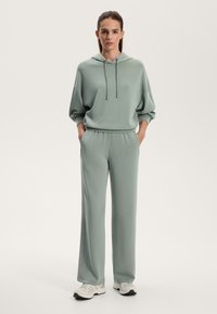 Világoszöld kapucnis pulóver zsinórral, túlméretezett szabásban, lazán illeszkedő melegítőnadrággal párosítva. Az anyag lágy és sima, fehér sportcipővel kiegészítve.