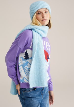 Gorro de punto azul claro, bufanda de canalé a juego y un suéter morado con un gráfico de montaña. Jeans de mezclilla casuales y una pose relajada.