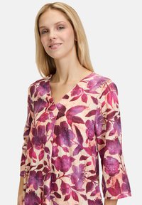 Blumen-Tunika-Kleid mit V-Ausschnitt, ¾-Ärmeln und einem lila und rosa Blumenmuster auf creamfarbenem Hintergrund, aus leichtem Stoff.