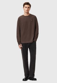 Felpa marrone a maniche lunghe con scollo rotondo, vestibilità rilassata e logo discreto sul petto, abbinata a jeans neri a gamba dritta e scarpe nere.