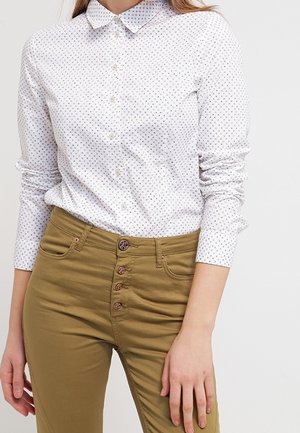 Vrouw die een wit langmouws overhemd met kleine stippen draagt en een beige broek met hoge taille en vier zichtbare knopen aan de voorkant.