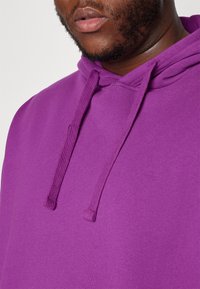 Violetti puuvillainen huppari, jossa on kiristysnauhallinen huppu, sileä pinta ja rento istuvuus, korostaen kankaan tasainen väri ja ompeleiden yksityiskohdat.