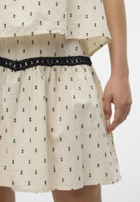 Robe couleur crème avec un motif à pois noirs, présentant une jupe froncée et une ceinture en dentelle noire en accent. Tissu léger avec une coupe décontractée.