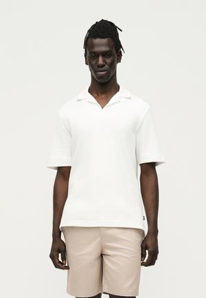 Άνδρας με πλεξούδες, ντυμένος με λευκό κοντομάνικο πουκάμισο με γιακά και μπεζ κοντά παντελόνια, στέκεται μπροστά σε ένα απλό φωτεινό φόντο.