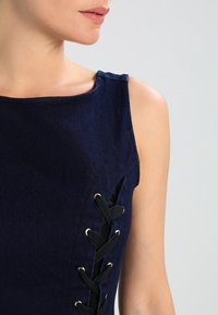 Kvinna som bär en ärmlös marinblå topp med svart snörning på sidan, visad från axlarna till mitten av överkroppen mot en ljus bakgrund.