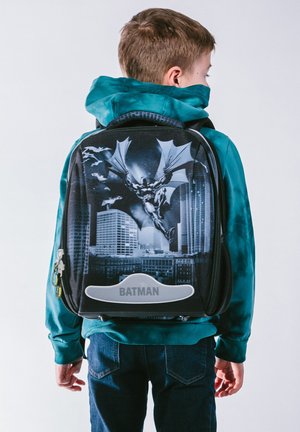 Zaino nero con grafica di Batman, dettagli argento e una texture liscia con zip. Un bambino indossa una felpa con cappuccio color teal.