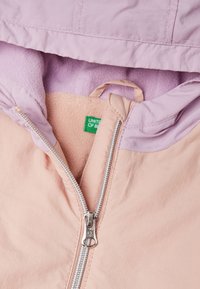 Giacca a vento rosa chiaro con una morbida fodera in pile viola, zip argentata e etichetta con la scritta "UNITED OF BOUNCE" all'interno.
