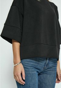 Black oversized short-sleeve top with a smooth texture and seam detailing, paired with blue denim jeans. Silver bracelet on wrist.