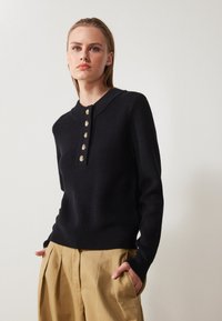 Svart ribbat stickat tröja med rund halsringning, med detaljer i guldtonade knappar fram. Ihopparad med beiga, veckade byxor.