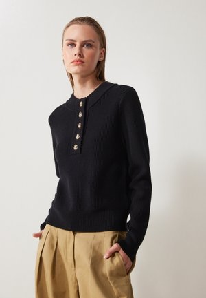 Un pull en tricot côtelé noir avec un col ras du cou, présentant des boutons dorés sur le devant. Associé à un pantalon beige plissé.