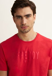 T-shirt rossa in cotone a maniche corte, con testo in rilievo "FREDDY TRAINING DEPT." di una tonalità leggermente più scura sulla parte anteriore.