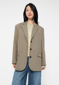 Junge Person mit glattem schwarzen Haar, die ein übergroßes taupefarbenes Blazer, eine hellgrüne Strickjacke, ein beiges Oberteil und lockere blaue Jeans trägt, vor einem weißen Hintergrund.
