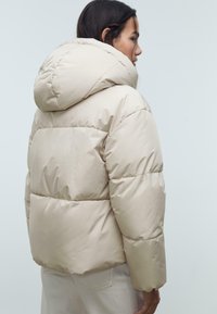 Bež puffer jakna s velikom kapuljačom, s kvadratnim uzorkom i bočnim džepovima. Glatka, sjajna tekstura, do koljena.