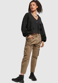 Kvinne med krøllete hår i en svart knappet kort genser, khaki cargo-bukser, svarte høye sneakers og et sølvkjede, poserer.