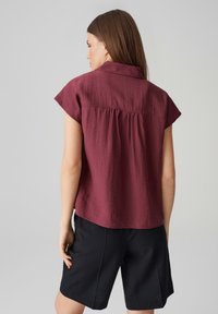 Bordeaux korte mouw blouse met een kraag, textuurstof en een geplooide achterkant, gecombineerd met zwarte shorts.