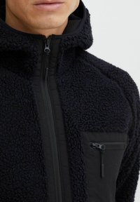 Chaqueta de forro polar negra con tejido texturizado, cremallera frontal y bolsillo en el pecho con cremallera. Presenta una capucha y un panel liso de contraste.