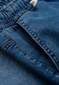 Tejido de mezclilla en azul oscuro, con una cintura fruncida, detalles de costura visibles y una textura suave. Apariencia ligeramente arrugada.