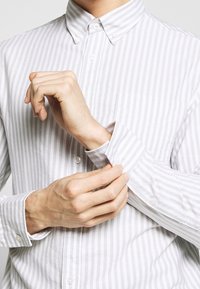 Světle šedá a bílá pruhovaná košile na knoflíky s ostrým límcem, nastavovacími manžetami a malou náprsní kapsou. Bavlněná tkanina, hladká textura.