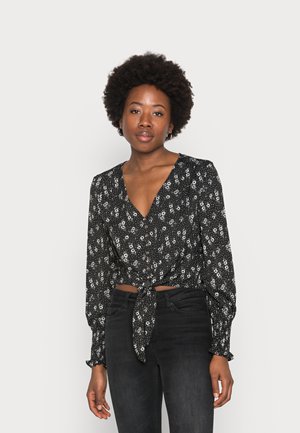 Top curto preto com padrão floral, mangas longas, decote em V, detalhe de laço na frente e acabamentos em botão. O padrão apresenta pequenas flores brancas sobre um fundo escuro.