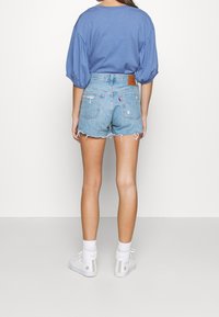 Camiseta oversized azul, pantalones cortos de mezclilla azul claro con dobladillos deshilachados y zapatillas altas blancas con acentos rosas. Etiqueta de cuero marrón en los pantalones cortos.