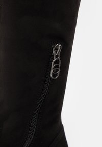 Bota de gamuza negra hasta la rodilla que cuenta con un cierre lateral y herrajes plateados, con una textura suave. Diseño estructurado con acentos mínimos.