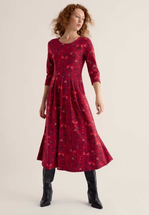 Femme aux cheveux bouclés portant une robe midi rouge à fleurs avec des manches trois-quarts et des bottes noires montantes jusqu'aux genoux, debout devant un fond uni.