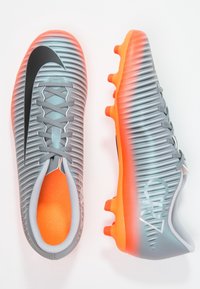 Šedé a oranžové fotbalové kopačky s texturovaným svrškem, vybavené černým logem Nike, dynamickými pruhy a oranžovými kolíky pro trakci.