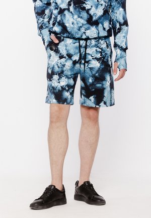 Mężczyzna w niebieskich i czarnych szortach tie-dye oraz pasującej bluzce z długim rękawem i czarnych trampkach, stoi z jedną ręką w kieszeni.
