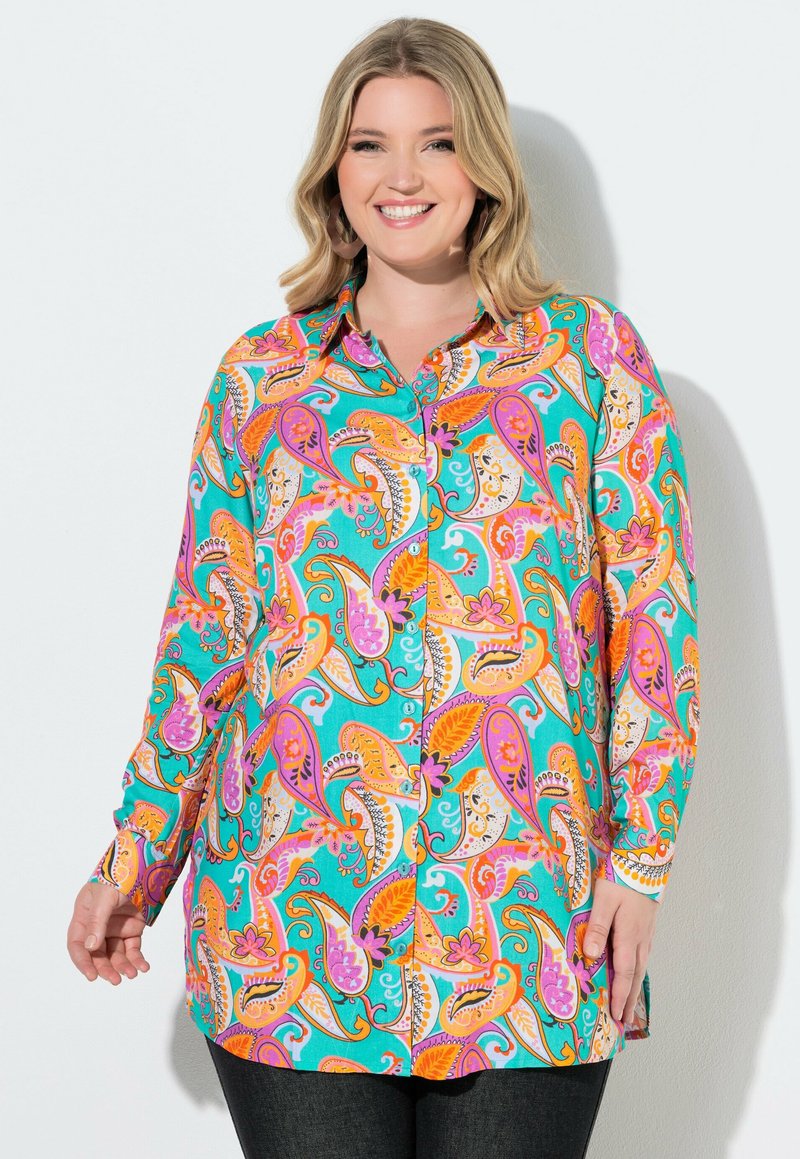 Mujer sonriente que lleva una blusa de manga larga con botones y estampados de cachemira en colores turquesa brillante, naranja, púrpura y blanco, de pie contra una pared blanca.