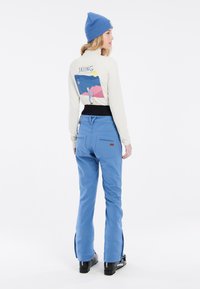 Blauwe skibroek met een hoge tailleband, zakken en ritsen bij de enkels. Beige shirt met lange mouwen heeft een kleurrafisch skitekening op de achterkant.