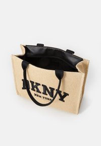 Sac cabas en toile beige avec texte de logo noir et bordure noire. Ouverture sur le dessus avec doublure intérieure noire et deux poignées noires. Design simple et rectangulaire.