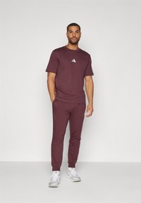 Burgundi színű sportöltözet, amely rövid ujjú pólóból áll, kis fehér logóval, és hozzá illő, tapered szabású melegítőnadrágból, mindkettő puha anyagból készült.