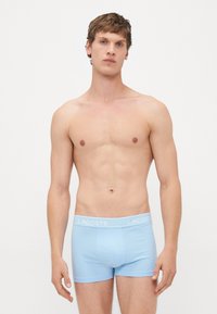 Joven de pie contra un fondo blanco, vistiendo calzoncillos boxer de Lacoste en azul claro, mirando al frente con una expresión neutral.