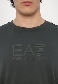 Tamsiai pilkas medvilnės marškinėliai su iškilniu guminiu logotipu juoda spalva, ant kurio užrašyti "EA7" ir "Emporio Armani". Klasikinio apvaliuko dizainas.
