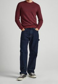 Röd och marinblå randig långärmad skjorta, mörkblå jeans, grå sneakers, uppvisad med händerna i fickorna, stående på en ljus bakgrund.