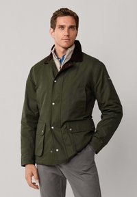 Chaqueta encerada verde oscuro con cuello de pana marrón, botones de presión frontales, dos bolsillos inferiores y un ajuste estructurado. Llevada sobre un suéter claro.