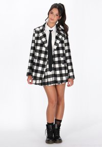 Černobíle kostkovaný dvouřadý blazer s klopami, kombinovaný s plisovanou sukní a černými šněrovacími botami, které dotvářejí celý outfit.