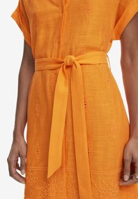 Vestito arancione ricamato con vita accentuata da una cintura. Presenta bottoni, maniche corte e motivi floreali lungo l'orlo per texture e dettaglio.