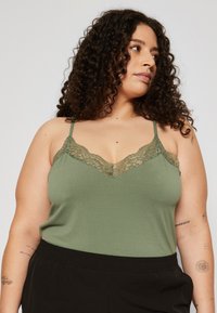 Olivgrünes Cami-Top mit Spitzenrand am Ausschnitt, dünnen verstellbaren Trägern und glatter Textur, kombiniert mit schwarzen Hosen mit elastischem Bund.