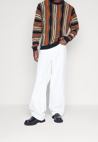 Der gestrickte Pullover zeichnet sich durch auffällige, mehrfarbige Streifen in Orange, Grün, Schwarz und Weiß aus; kombiniert mit lockeren weißen Hosen und schwarzen Schuhen.