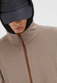 Brun teksturert jakke med glidelås og høy krage, lagd under en svart hettecap. Designet er sport og uformelt.