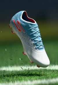 Light blue football boot with white and pink accents, textured upper, and laces. Cleats visible, suspended above grass.