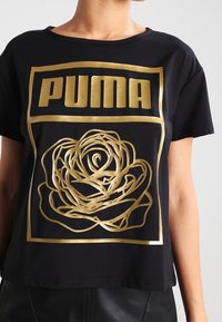 Camiseta negra con un logo dorado de "PUMA" y un diseño de rosa. La camiseta tiene un corte relajado y una textura de tela suave.
