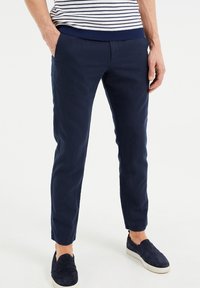 Man i marinblå slim-fit byxor och matchande slip-on skor, med händerna i fickorna, som står mot en vit bakgrund.