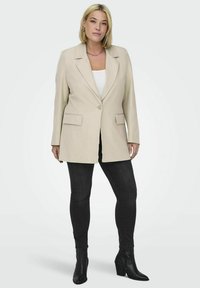 Béžový oversized blejzr s dvěma předními kapsami a zapínáním na jeden knoflík, v kombinaci s bílým topem a úzkými černými džíny, na jednobarevném pozadí.