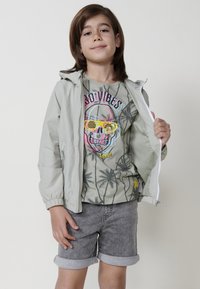 Giacca a vento verde chiaro con polsini elasticizzati, t-shirt grigia con grafica di un teschio con occhiali da sole colorati, pantaloni corti di denim con orlo arrotolato.