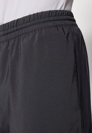 Gros plan sur la zone de la taille présentant une ceinture élastique noire et un tissu lisse d'un pantalon léger porté par une personne vêtue d'un haut blanc texturé.