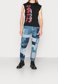 Czarna bezrękawnik z czerwonym nadrukiem graficznym, w połączeniu z niebieskimi dżinsami w stylu patchwork z różnymi odcieniami i postarzanym wykończeniem. Czarne buty.