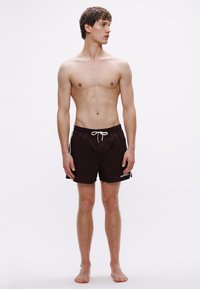 Bañador corto color granate con cintura de cordón, con detalle de logo lateral. Modelo descalzo, mostrando un diseño minimalista.